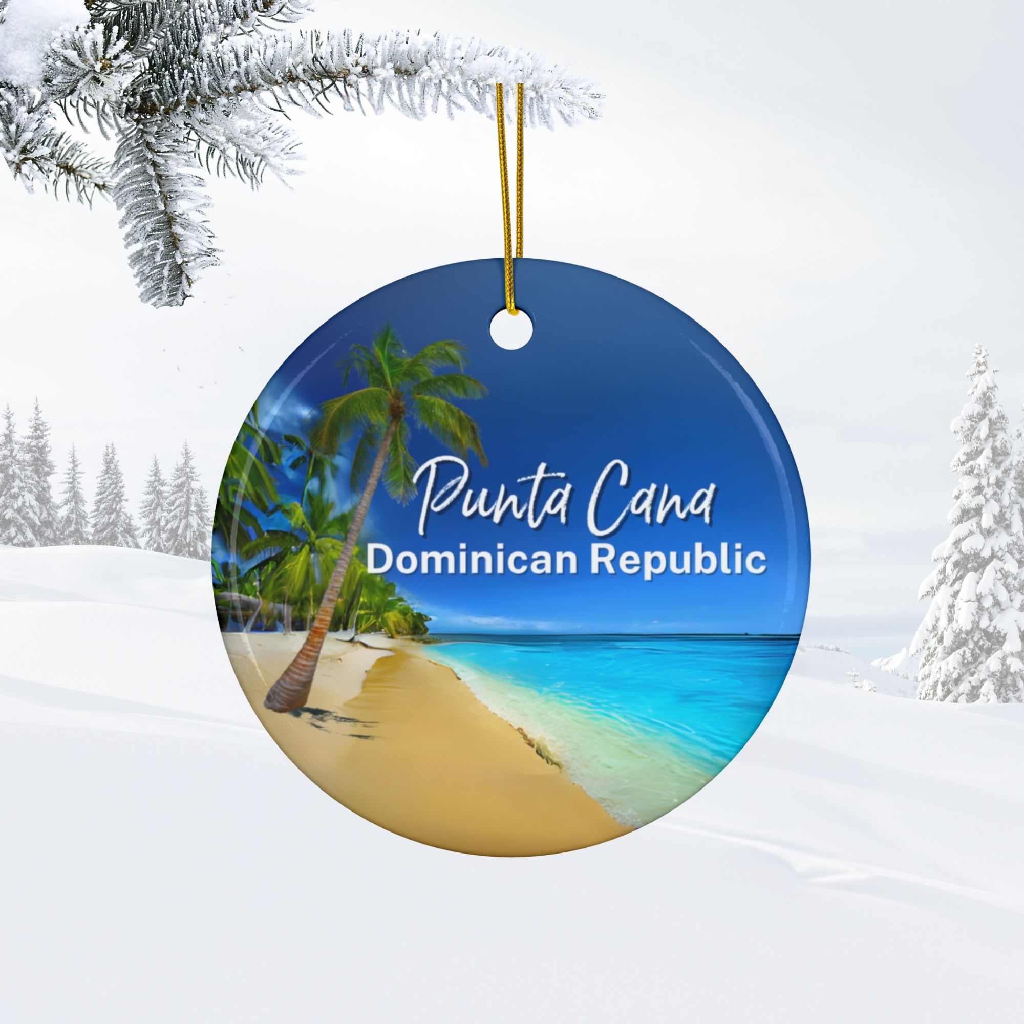 Double-sided Punta Cana ornament featuring a tropical beach scene and palm trees, labeled Punta Cana Dominican Republic, hangs from a gold string in a snowy winter landscape with pine branches and snow-covered trees.
